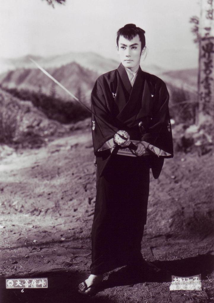 A young man dressed in traditional Japanese clothing stands confidently with a sword, set against a mountainous backdrop.
