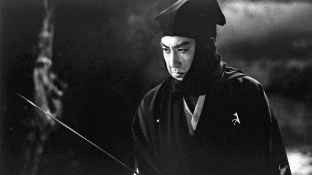 A black and white image of a focused male character dressed as a ninja, holding a weapon and appearing tense in a dramatic outdoor setting.