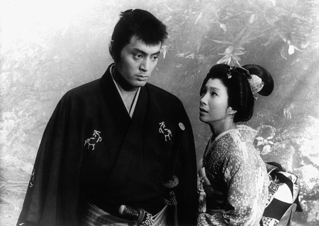 A black and white image of a man in traditional Japanese clothing, looking serious, standing next to a woman in a patterned kimono, who appears to be speaking to him with an expression of concern.