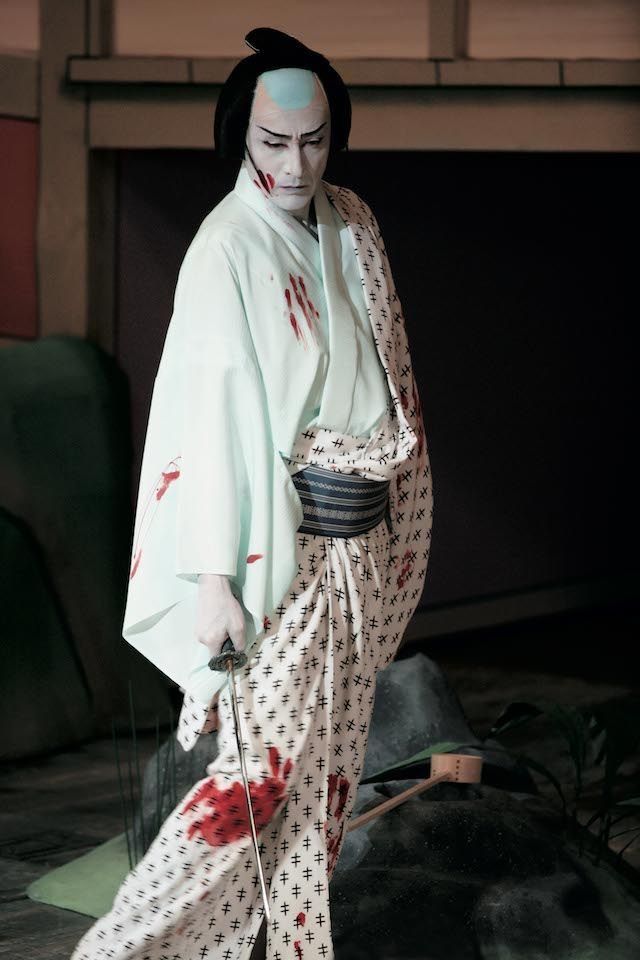 A performer dressed in a traditional kimono with bloodstains, holding a sword, depicted in a dramatic pose.