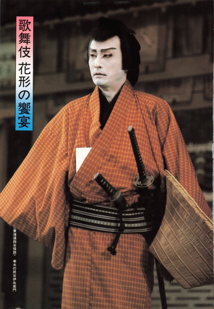 A traditional Japanese performer dressed in an orange kimono with a black and white obi, holding a fan and displaying two sheathed swords, standing against a blurred background.
