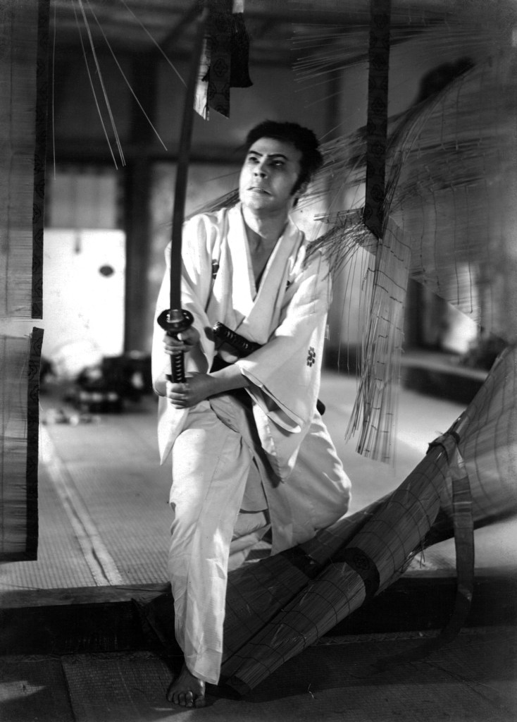 A man in traditional Japanese clothing holds a katana, preparing to strike, with a focused expression in a dimly lit room.