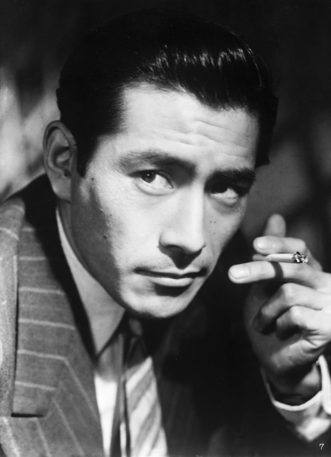 Black and white portrait of a man in a pinstriped suit, looking intently at the camera while holding a cigarette between his fingers.