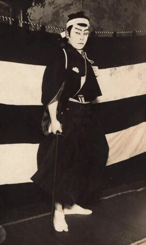 A historical black and white photo of a Japanese actor in traditional attire, standing with a sword in front of a striped backdrop.