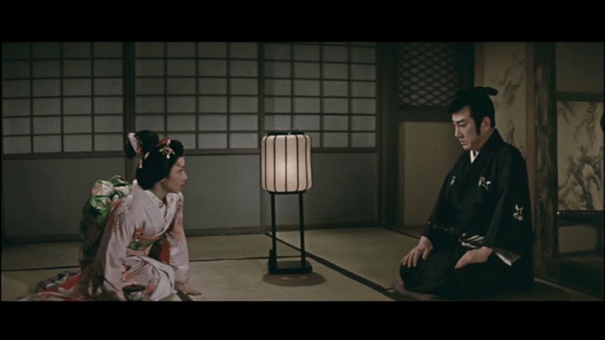 A scene in a traditional Japanese room featuring a man in a black kimono sitting on the floor and a woman in a pink kimono kneeling nearby. A soft lantern is illuminated between them.