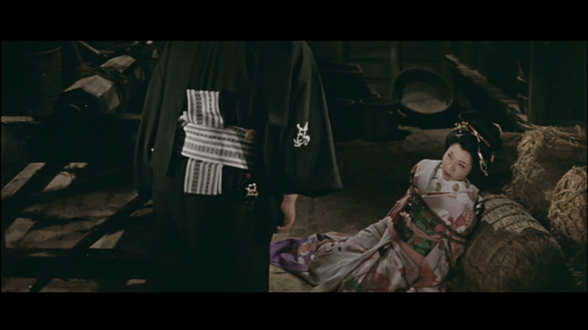 A dramatic scene featuring a person in traditional Japanese clothing standing next to another person seated on the ground, dressed in a colorful kimono, with straw bales in the background.