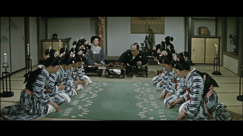 A traditional Japanese room with several women in striped kimonos seated on the floor, participating in a game. In the background, a man in traditional attire is observing while another woman is preparing food on a table.