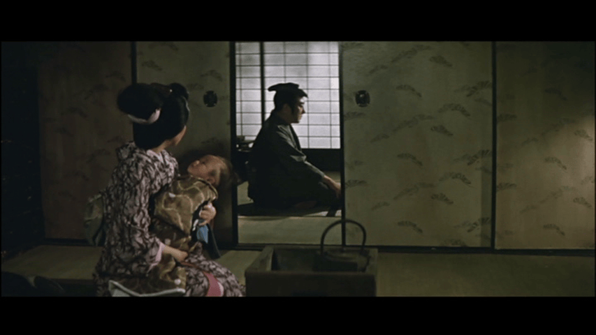 A woman in traditional attire holds a baby while looking towards a seated man in a dimly lit room, with paper sliding doors and a tranquil atmosphere.