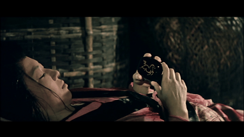 A woman lying down, holding a decorative black object with intricate designs, in a dimly lit, rustic setting.