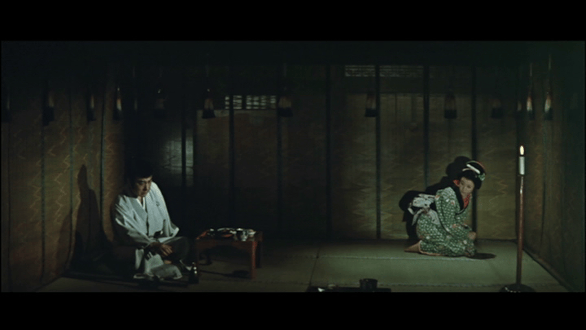 A man in traditional clothing sits on the floor, while a woman in a patterned kimono crouches nearby in a dimly lit room with wooden walls.