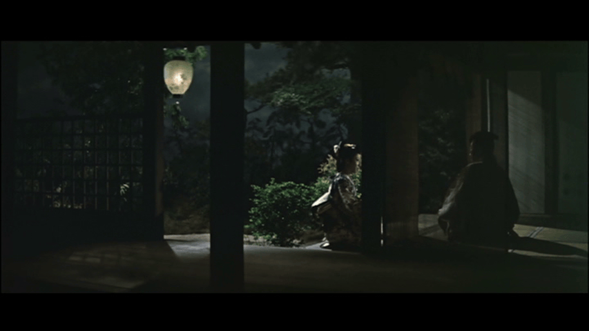 A dimly lit scene featuring a woman in traditional attire sitting on a porch, with a lantern glowing softly above her, while a man sits to the side, set against a backdrop of trees at night.