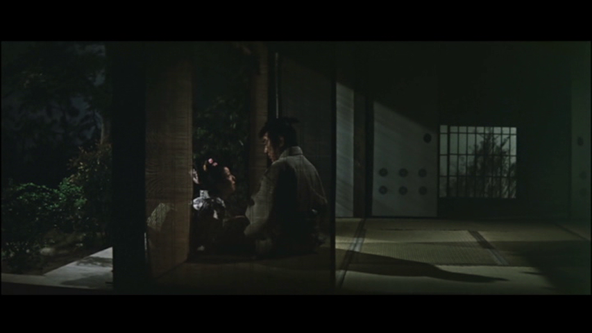 A dimly lit scene featuring two figures, a man and a woman, sitting together near an open wooden door. The atmosphere is intimate, with a garden visible outside.